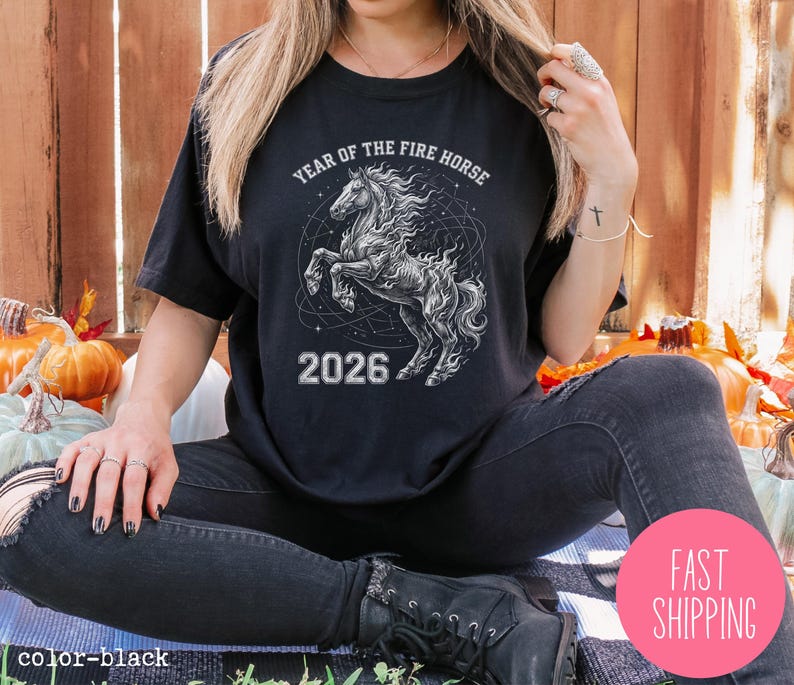 K&ouml;nnte beinhalten: Schwarzes T-Shirt mit einer detaillierten Illustration eines sich aufb&auml;umenden Pferdes, der Text lautet "YEAR OF THE FIRE HORSE" und "2026". Das Design ist in Wei&szlig;- und Graut&ouml;nen gehalten. Das T-Shirt ist schwarz.