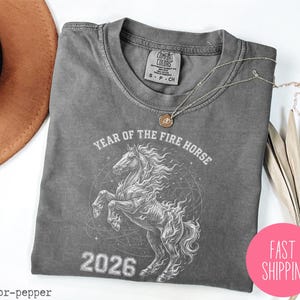 K&ouml;nnte beinhalten: Ein graues T-Shirt mit einer Grafik eines sich aufb&auml;umenden Pferdes, dem Text "YEAR OF THE FIRE HORSE 2026" und einem Comfort Colors-Etikett. Ein rosa Kreis mit "FAST SHIPPING" befindet sich in der unteren rechten Ecke.