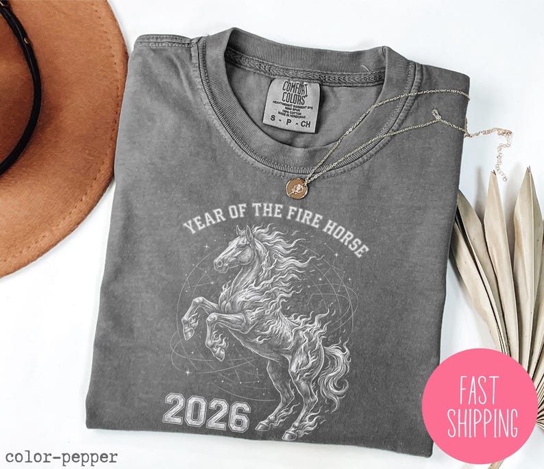 K&ouml;nnte beinhalten: Ein graues T-Shirt mit einer Grafik eines sich aufb&auml;umenden Pferdes, dem Text "YEAR OF THE FIRE HORSE 2026" und einem Comfort Colors-Etikett. Ein rosa Kreis mit "FAST SHIPPING" befindet sich in der unteren rechten Ecke.