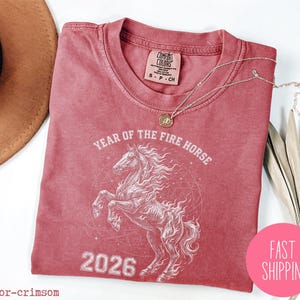 K&ouml;nnte beinhalten: Karmesinrotes T-Shirt mit einer wei&szlig;en Pferde-Grafik und dem Text "YEAR OF THE FIRE HORSE 2026". Das Shirt hat einen Rundhalsausschnitt und kurze &Auml;rmel. Eine goldene Halskette liegt &uuml;ber dem Shirt. Ein rosa Kreis mit "FAST SHIPPING" befindet sich unten rechts.