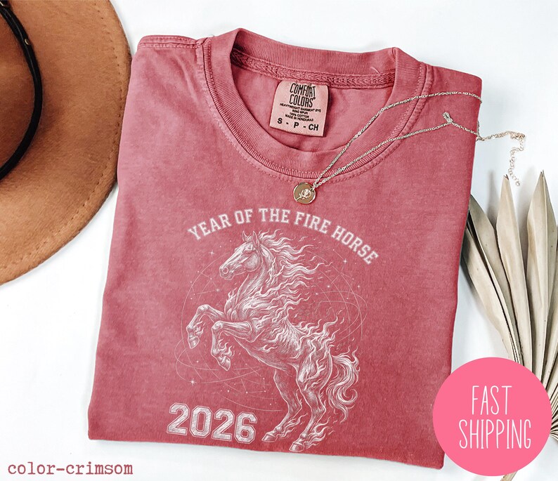 K&ouml;nnte beinhalten: Karmesinrotes T-Shirt mit einer wei&szlig;en Pferde-Grafik und dem Text "YEAR OF THE FIRE HORSE 2026". Das Shirt hat einen Rundhalsausschnitt und kurze &Auml;rmel. Eine goldene Halskette liegt &uuml;ber dem Shirt. Ein rosa Kreis mit "FAST SHIPPING" befindet sich unten rechts.