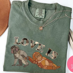 May include: A moss green t-shirt with the words "LOVE IS" in a patchwork design, featuring two monkey illustrations. A gold necklace is draped across the shirt. The shirt has a Comfort Colors label.