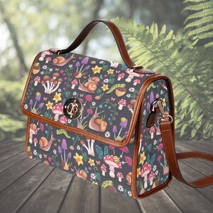 May include: A dark grey satchel bag featuring a pattern of mushrooms, snails, flowers, and butterflies. It has brown leather trim, a top handle, and a detachable shoulder strap. The clasp is gold-coloured.