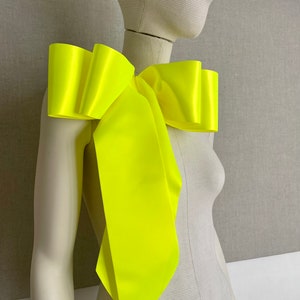 May include: A vibrant neon yellow bow accessory is displayed on a mannequin. The bow, made from a glossy fabric, has a voluminous, ruffled design across the shoulder. Long, flowing tails cascade down, adding a dramatic effect to the fashion item.