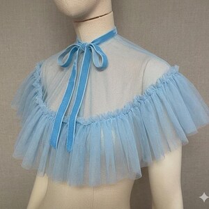 May include: A light blue tulle cape with a ruffled edge and a velvet tie. The cape is displayed on a mannequin. The neck tie is a matching light blue velvet.