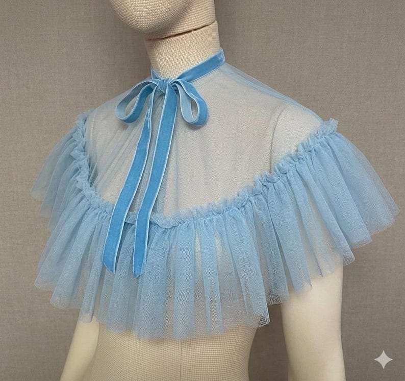 May include: A light blue tulle cape with a ruffled edge and a velvet tie. The cape is displayed on a mannequin. The neck tie is a matching light blue velvet.