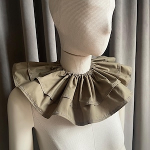 Puede incluir: Un cuello con volantes de color beige en un maniquí. El cuello tiene múltiples capas de tela, creando un efecto voluminoso. El maniquí se exhibe sobre un fondo gris neutro.