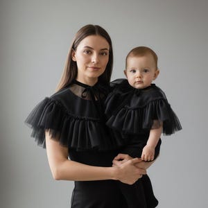 May include: A woman and a baby wearing matching black outfits. The outfits include a black dress and a ruffled tulle cape. The woman is holding the baby. The background is a solid gray.