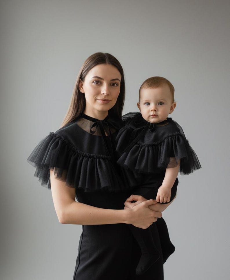 May include: A woman and a baby wearing matching black outfits. The outfits include a black dress and a ruffled tulle cape. The woman is holding the baby. The background is a solid gray.