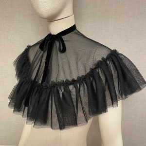 May include: A black tulle collar with a velvet ribbon tie. The collar features a ruffled edge and is displayed on a mannequin. The sheer fabric creates a delicate and elegant look, suitable for formal wear or costume.