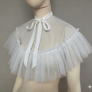May include: A white tulle collar with a ruffled edge and a satin ribbon tie. The collar is designed to be worn around the neck and shoulders. The fabric is sheer and delicate, creating a soft, ethereal look.