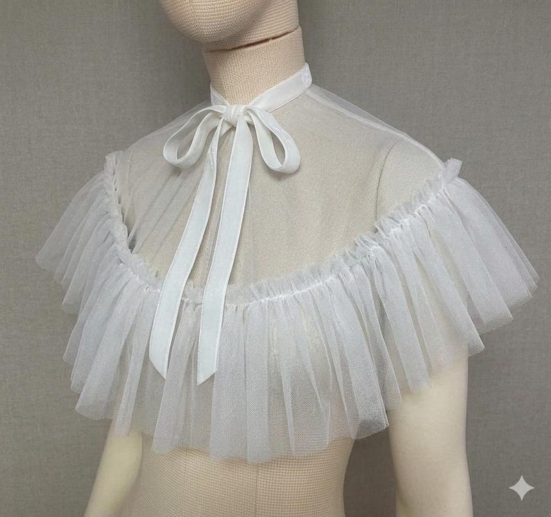 May include: A white tulle collar with a ruffled edge and a satin ribbon tie. The collar is designed to be worn around the neck and shoulders. The fabric is sheer and delicate, creating a soft, ethereal look.