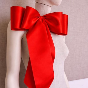 Könnte beinhalten: Ein großer, leuchtend roter Satin-Schleife, die über der Schulter einer Schaufensterpuppe drapiert ist. Die Schleife hat mehrere Schlaufen und lange, fließende Enden, wodurch ein dramatisches und elegantes Accessoire entsteht. Die Schleife ist ein auffälliges Statement-Piece.