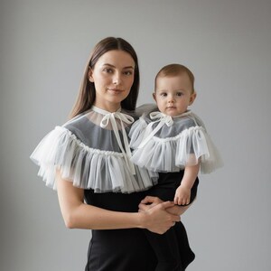 May include: A woman and a baby wearing matching black outfits with white tulle capes. The capes have a ruffled edge and a bow at the neckline. The woman is holding the baby, both looking at the camera.