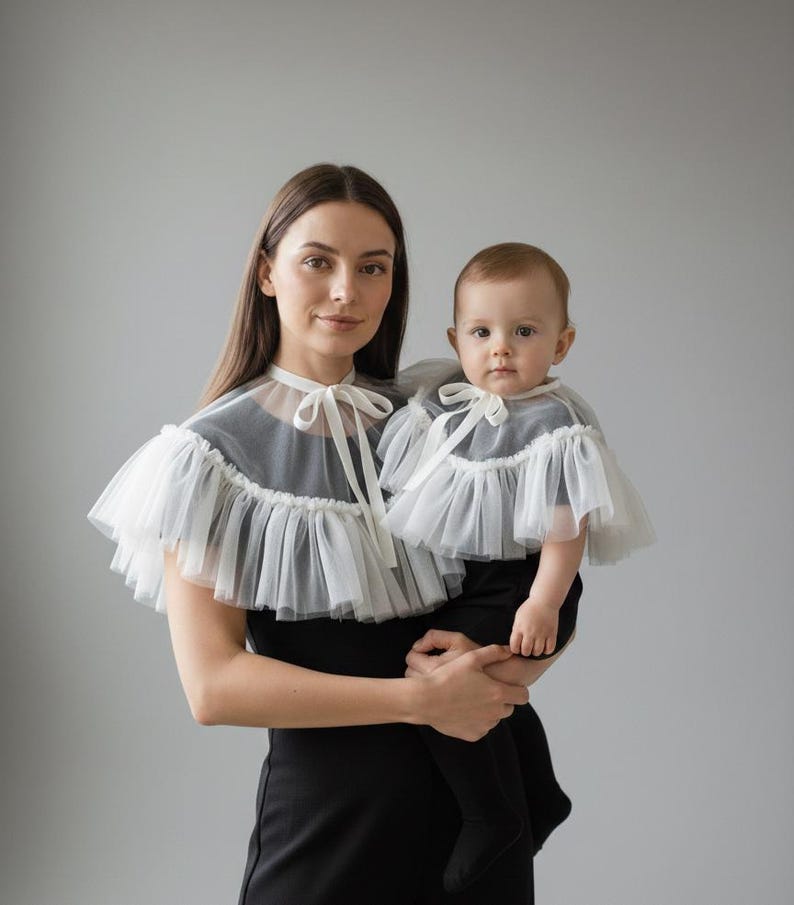 May include: A woman and a baby wearing matching black outfits with white tulle capes. The capes have a ruffled edge and a bow at the neckline. The woman is holding the baby, both looking at the camera.
