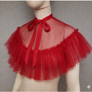 May include: A red tulle cape with a ruffled edge and a high collar. The cape features a satin ribbon tie at the neck. The sheer fabric creates a delicate and elegant look, suitable for special occasions or costume wear.