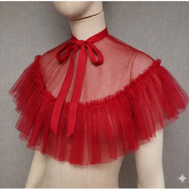 May include: A red tulle cape with a ruffled edge and a high collar. The cape features a satin ribbon tie at the neck. The sheer fabric creates a delicate and elegant look, suitable for special occasions or costume wear.