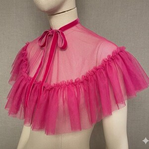 May include: A vibrant pink tulle capelet with a ruffled edge and a velvet ribbon tie. The capelet is sheer and layered, with a high neckline and a delicate, feminine design. The color is a bright, eye-catching pink.