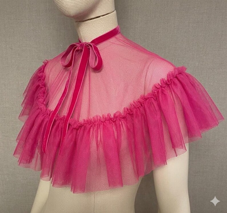 May include: A vibrant pink tulle capelet with a ruffled edge and a velvet ribbon tie. The capelet is sheer and layered, with a high neckline and a delicate, feminine design. The color is a bright, eye-catching pink.