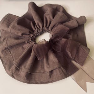 May include: A brown fabric hair scrunchie with a large brown tulle bow. The scrunchie is made of multiple layers of fabric and has a gathered look.