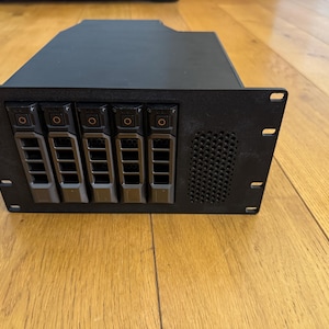Könnte beinhalten: Schwarzer Server-Rack mit fünf Laufwerkschächten und einem Lüftungsgitter. Jeder Laufwerkschacht hat einen kleinen schwarzen Knopf.
