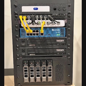 Puede incluir: Un rack de servidor negro lleno de hardware de red. Incluye un dispositivo blanco, un Palo Alto PA-220 azul, dos dispositivos Lenovo ThinkCentre y varias bahías de disco duro. Cables amarillos y negros conectan los dispositivos.