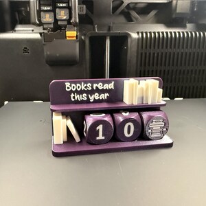 May include: A purple 3D-printed book counter with the text "Books read this year." The counter displays the number "10" with dice-shaped blocks. The top shelf holds small, rectangular book-shaped blocks.