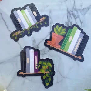 3 subtle pride stickers. Agender flag with black, grey, white, green, white, grey, black books and vines. Aromantic flag, with dark green, light green, white, grey and black books with a cactus. Asexual flag, black, grey, white, purple books w/ plant