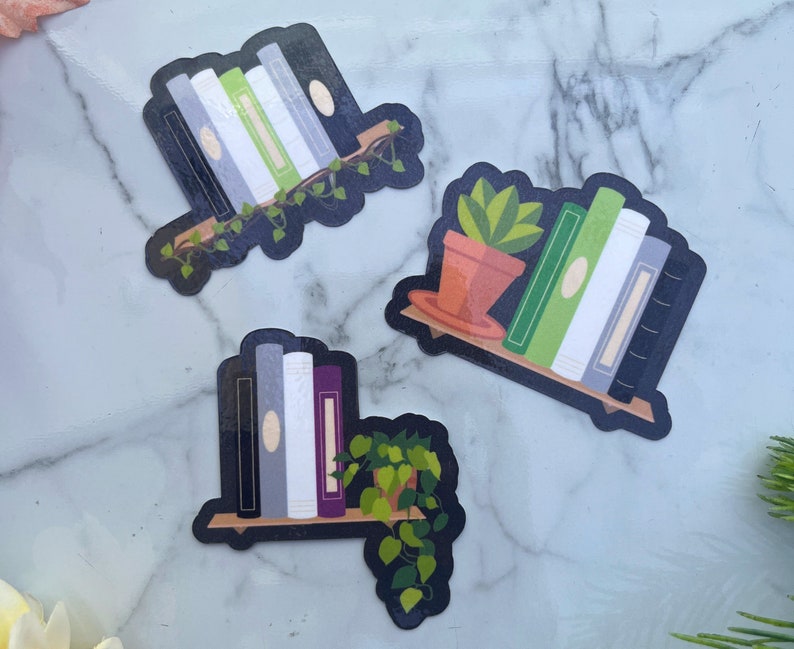 3 subtle pride stickers. Agender flag with black, grey, white, green, white, grey, black books and vines. Aromantic flag, with dark green, light green, white, grey and black books with a cactus. Asexual flag, black, grey, white, purple books w/ plant