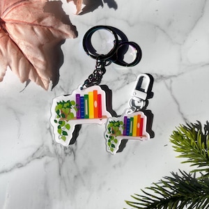 Könnte beinhalten: Zwei regenbogenfarbene schlüsselanhänger in Buchform mit einer kleinen Topfpflanze, die von dem Regal hängt. Die Schlüsselanhänger sind aus klarem Acryl und haben ein weißes Regal mit einem regenbogenfarbenen Stapel Bücher.