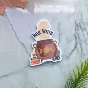 May include: A sticker featuring a brown cauldron with three crescent moons, steam rising, and the words "BASIC WITCH" in a curved font. A black cat sits beside the cauldron, with books and a cup of coffee. The sticker has a white border.