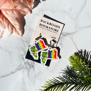 May include: Rainbow-colored book earrings with green vines, hanging from silver hooks. The earrings are on a white surface with a business card that reads "Backroads Bookstore, A Place for Bookish Merch".