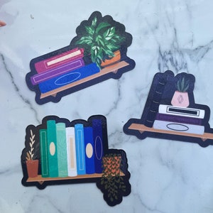 3 subtle pride stickers. Bisexual, horizontal pink, purple and blue books with a plant. Demisexual, upright black, horizontal white, purple and grey books with a succulent on top. Gay, range of teal to white to blue books with a plant on both sides.