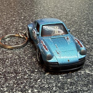 May include: A light blue metallic toy car keychain featuring a Porsche design with visible paint wear and rust-colored markings on the hood and sides. The miniature vehicle includes a silver metal keyring attached to the rear, black bumpers, and clear windows.