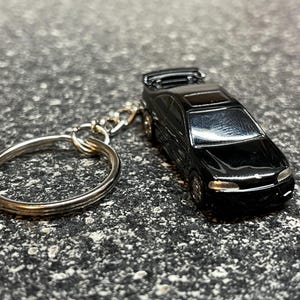 May include: A black miniature car keychain with a silver key ring. The car has a spoiler and detailed wheels. The keychain is on a textured gray surface.