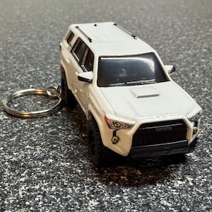 May include: A white Toyota 4Runner model keychain. The miniature SUV has black tires, a black front grill, and a silver key ring. The word "TOYOTA" is visible on the front.