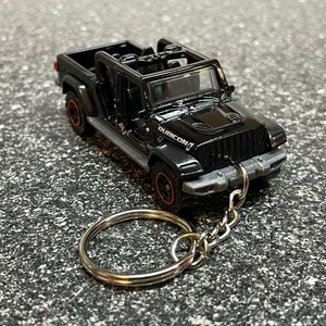 May include: Black Jeep Rubicon toy car keychain. The miniature vehicle has red-rimmed wheels and a silver keychain ring. The word "RUBICON" is visible on the hood. The toy car is on a textured surface.