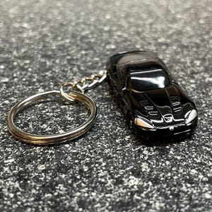 May include: A black die-cast car keychain. The miniature car has a glossy finish, silver accents, and a detailed design. The keychain includes a silver split ring and chain. The car measures approximately 3 inches in length.