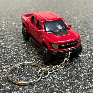 May include: Red Ford F-150 Raptor pickup truck keychain. The truck has a black front bumper and black wheels. The keychain has a silver metal ring and chain.