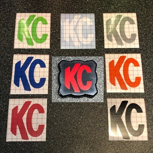 May include: Nine different versions of a "KC" logo sticker. The stickers are on a white background with a grid pattern. The logos are in various colors, including green, white, gray, blue, red, orange, and black.