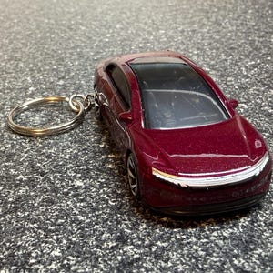 May include: A burgundy-colored car keychain. The miniature car has a sleek design with a black roof and silver accents. The keychain ring is silver and attached to the car's roof.
