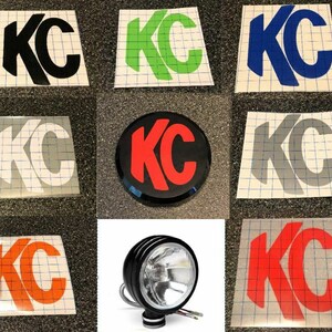 May include: Assortment of KC logo decals in various colors, including black, green, blue, white, red, orange, and gray. The decals are on a white background with a grid pattern. There is also a black round KC logo with a red center. A black KC off-road light is also included.