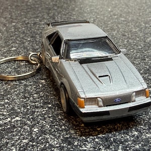 May include: A silver die-cast model car keychain. The miniature car features a detailed design with a spoiler, hood vent, and a Ford logo. The keychain ring is silver.