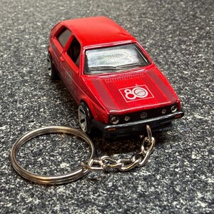 May include: A red die-cast model car keychain. The car has black trim, silver wheels, and a logo on the hood that says "80". The keychain has a silver ring and chain.