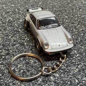May include: Silver die-cast model car keychain. The miniature car features a detailed design with a spoiler and is attached to a metal chain and keyring. The car measures approximately 3 inches in length.