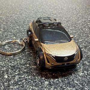 May include: A gold and black miniature car keychain. The car has a detailed design with black wheels, a black roof, and a silver key ring. The car's front features a Nissan logo.