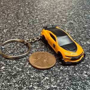 May include: A yellow and black toy car keychain featuring a small black Autobot logo on the side door. The car is positioned next to a copper-colored United States one-cent penny, which serves as a scale reference for the size of the vehicle.
