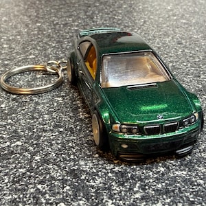 May include: A green miniature BMW M3 car keychain. The toy car has a metallic finish, black accents, and a silver key ring. The car's interior is visible through the windows. The keychain is on a textured surface.