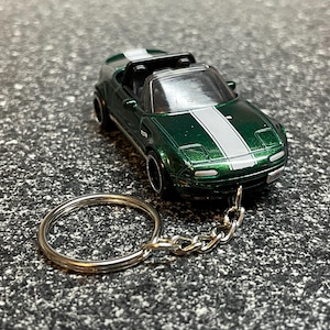 May include: A miniature green convertible car keychain with a silver racing stripe down the center. The car has black wheels and a silver keychain ring and chain. The car is a toy or model.