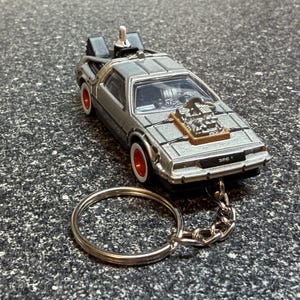 May include: A silver DeLorean keychain with red wheels and a silver chain. The miniature car has a detailed engine and the text "DMC" on the front. The keychain is on a textured gray surface.